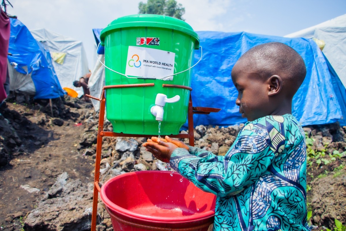 A young boy washes his hands using a handwashing station and tents can be seen behind him