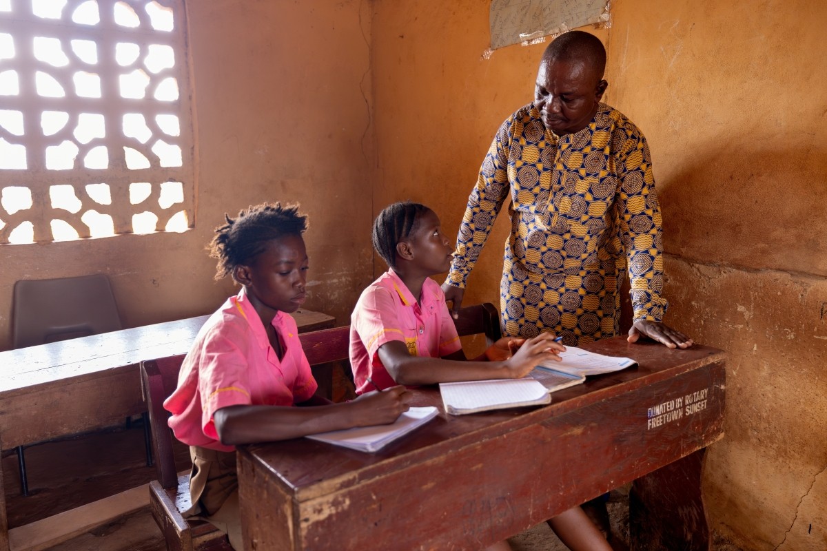 Male teacher stands beside two female students at desk, helping them with their work.